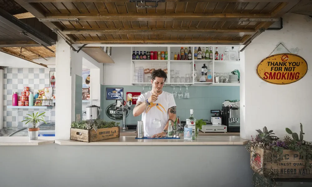 Una persona dietro il bancone di un bar prepara una bevanda, circondata da decorazioni e piante.