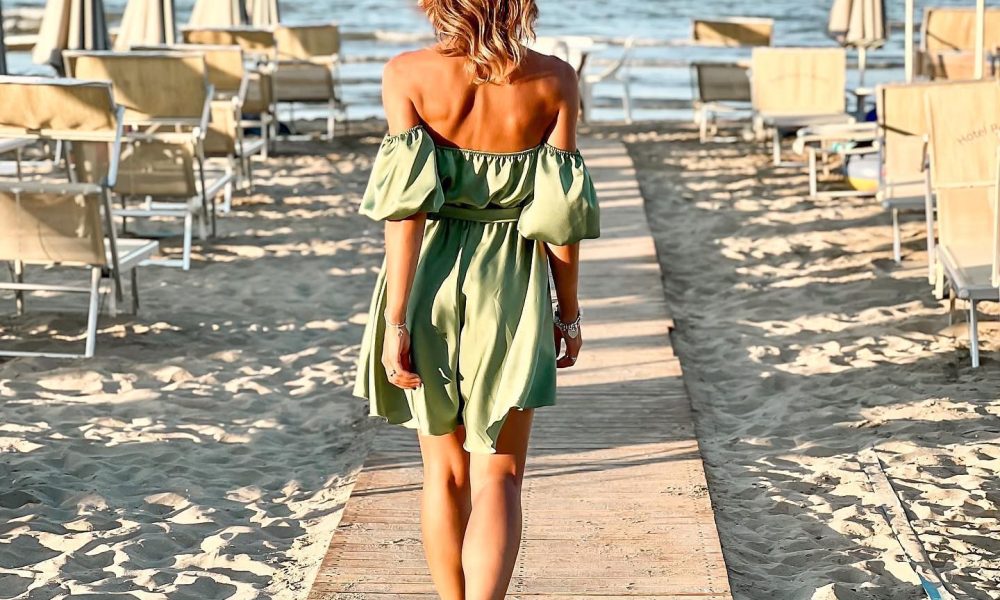 Une femme en robe verte marche sur une plage en direction de la mer par temps ensoleillé.